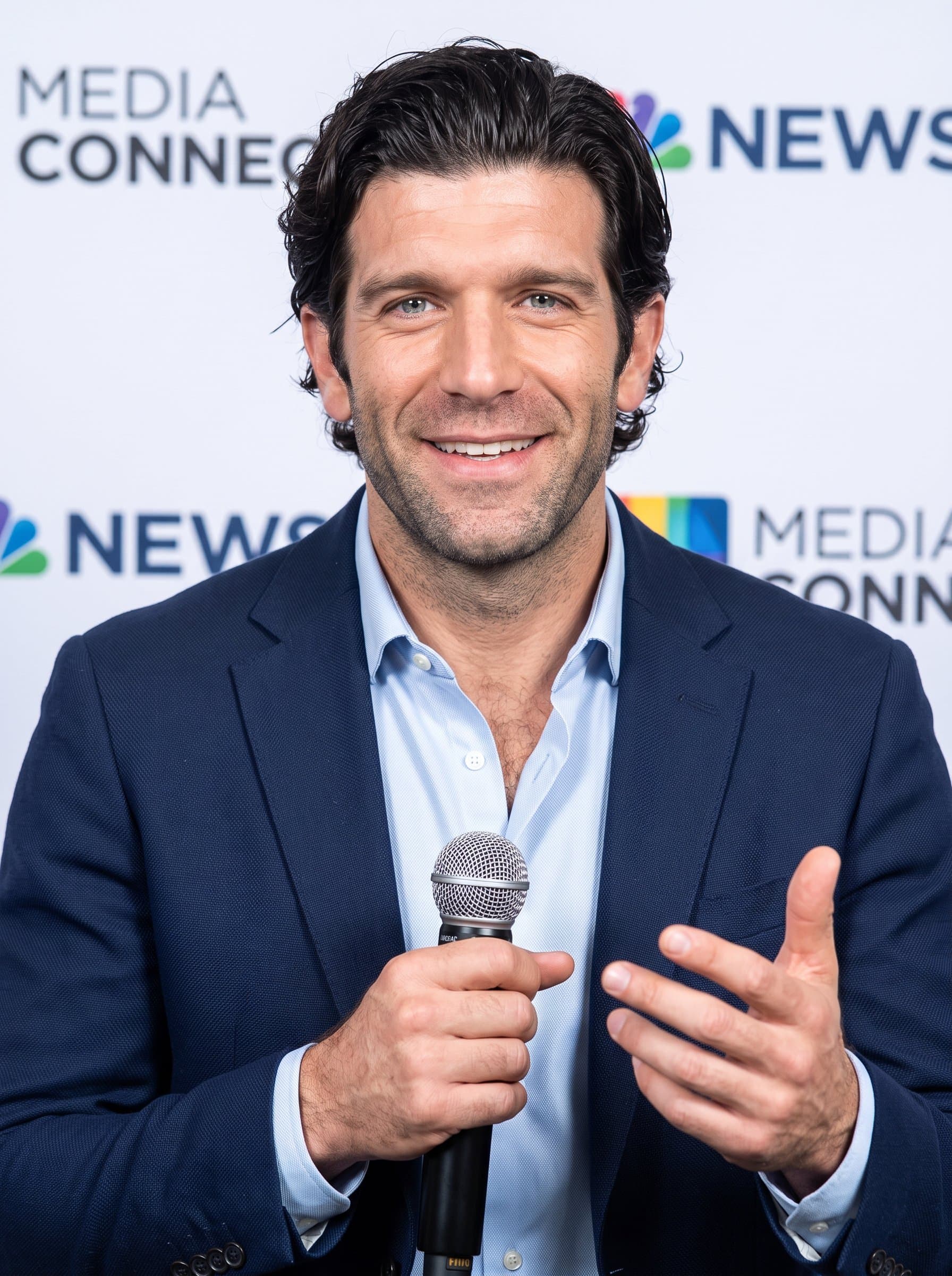 : Public relations professional headshot, highlighting a communicative man in stylish business wear, with a media-centric backdrop, embodying connectivity and charisma for a Glassdoor profile.