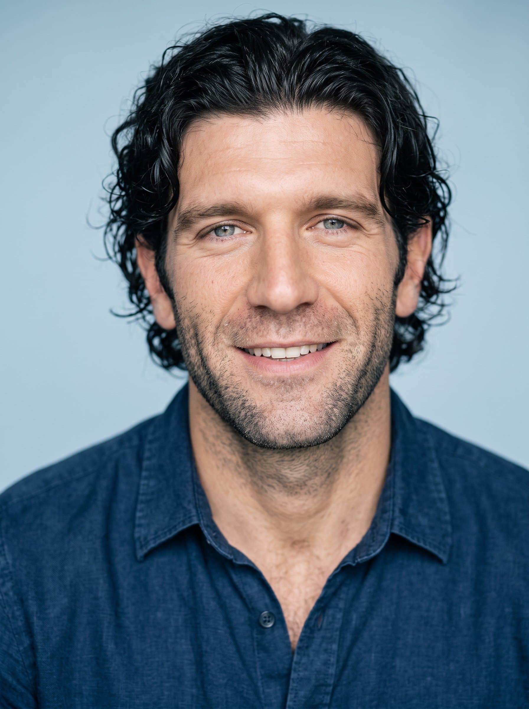 Capture in a headshot, presenting an inviting smile, dressed in a casual-collared shirt, set against a neutral soft light blue backdrop, enhanced by gentle, evenly distributed lighting.