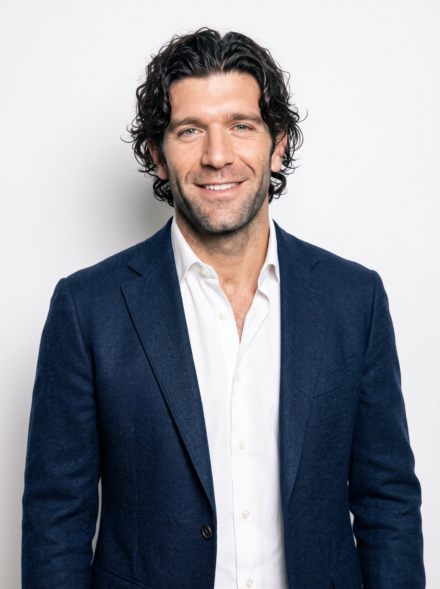 An image of featuring an man with curly hair, sporting a fitted blazer and open-collared shirt. Their poised posture and warm smile are set against a clean and simple white backdrop.