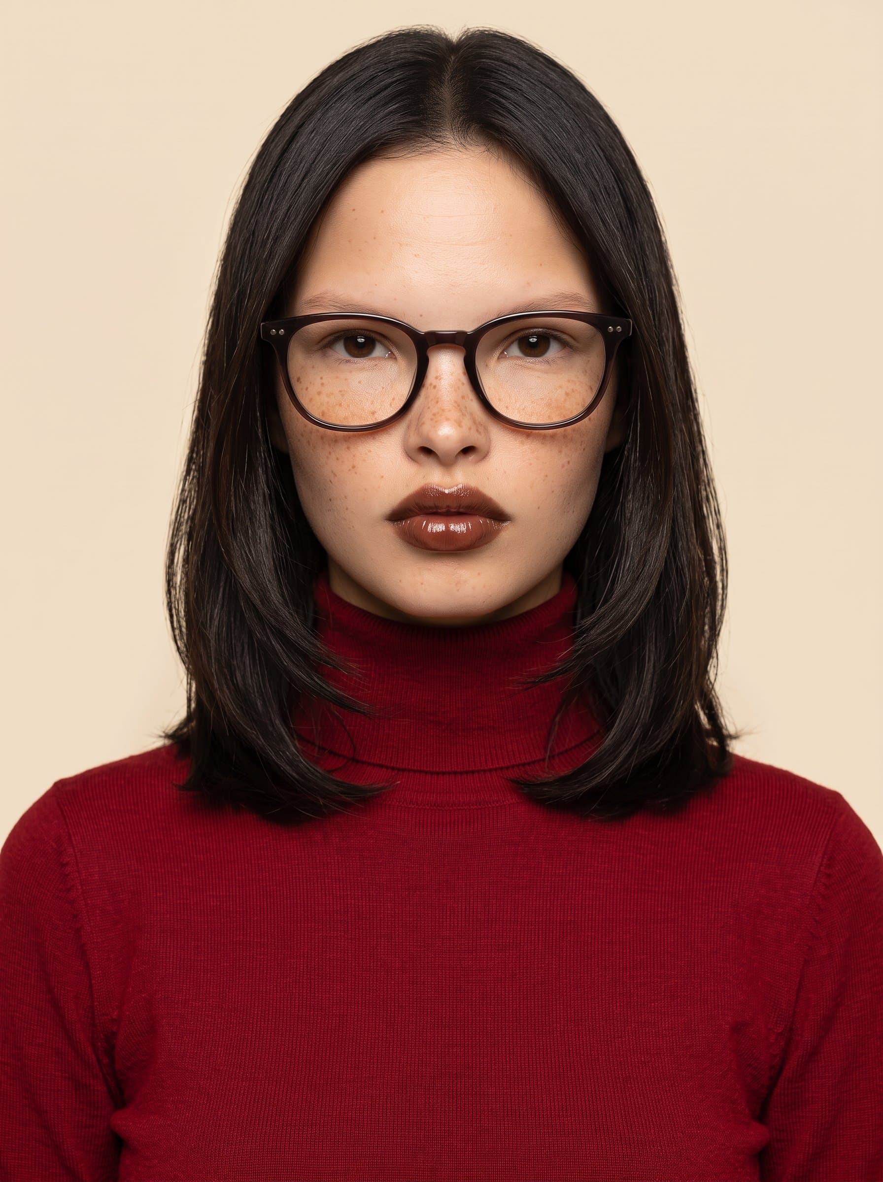 Under an woman with medium-length hair and glasses is pictured, wearing a red turtleneck sweater. A solid cream background adds to the image's professional yet warm feel.