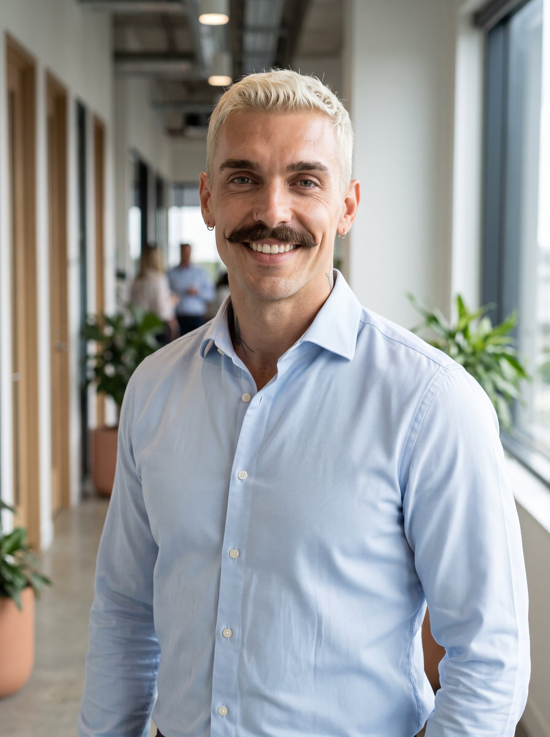 Show a classic professional in a muted pastel shirt, subtly illuminated by natural light. A blurred office corridor sets the scene, projecting professionalism and approachability.