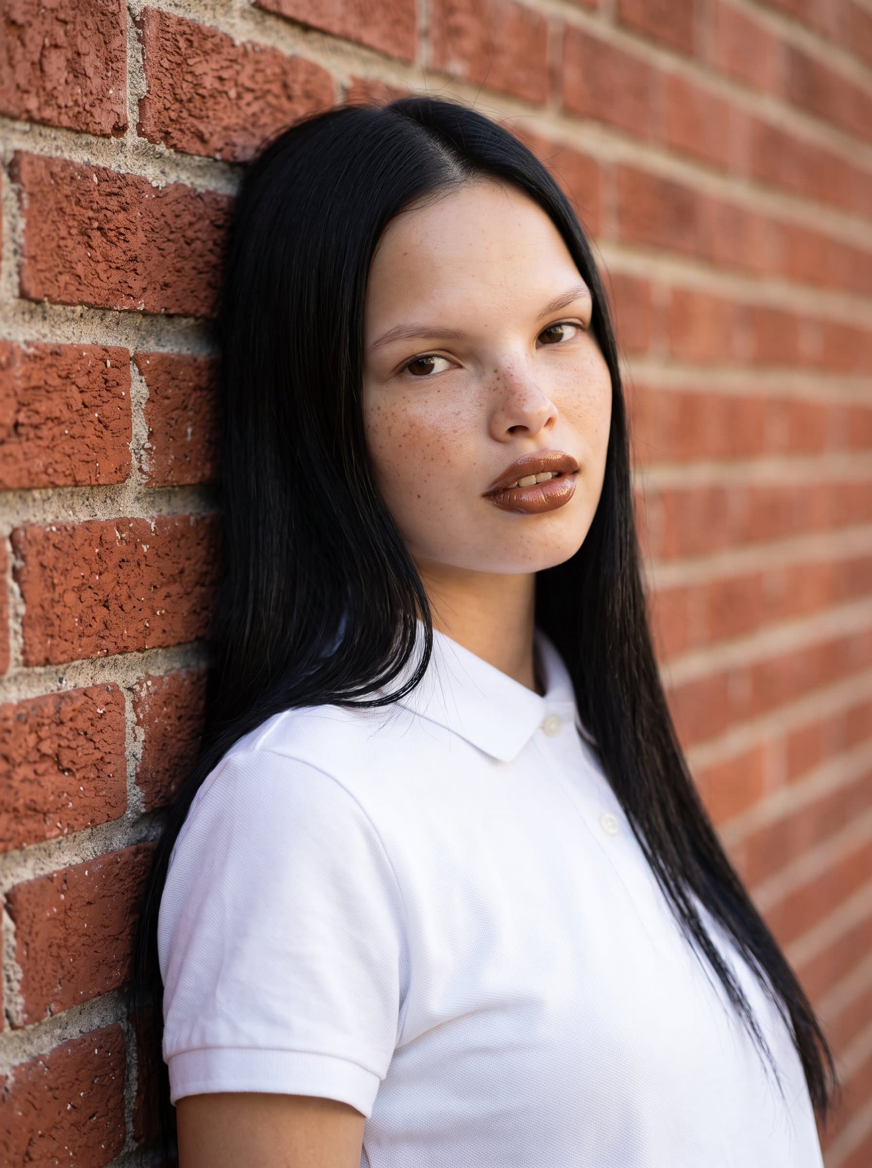 leans casually against a textured brick wall, wearing a simple collared shirt. The lighting creates soft shadows, adding depth to the headshot while maintaining a professional, inviting look.