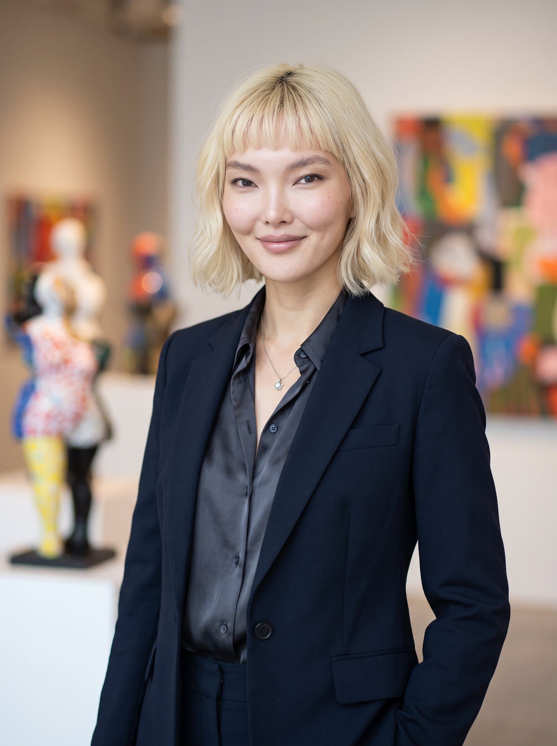 with a gentle, confident smile, stands before a softly blurred modern art installation. The professional attire and neutral lighting convey creativity and approachability in a balanced, professional manner.